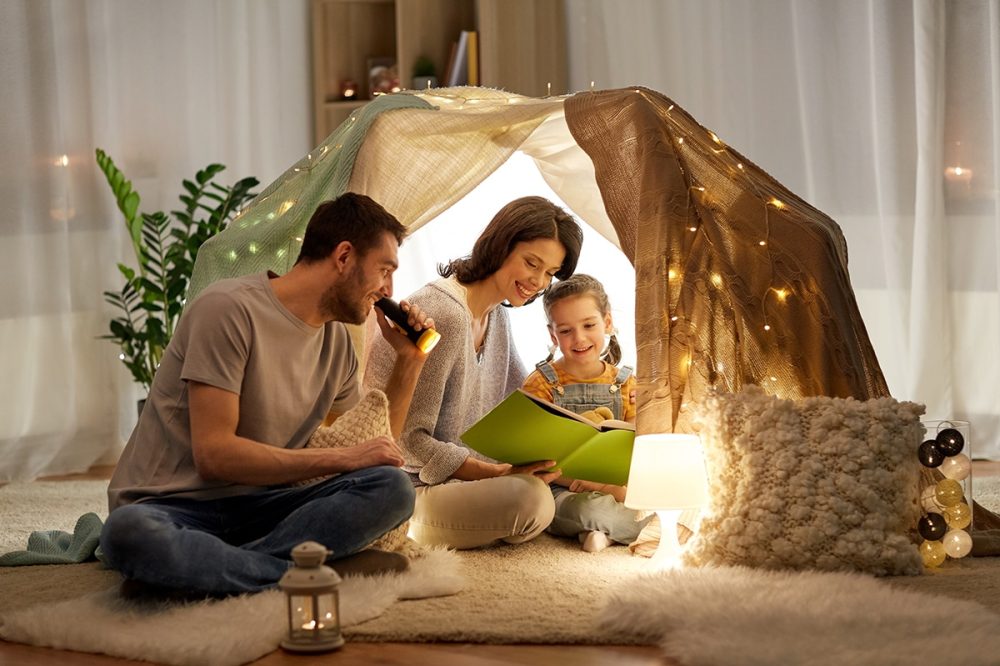 family, hygge and people concept - happy mother, father and little daughter reading book with torch light in kids tent at night at home